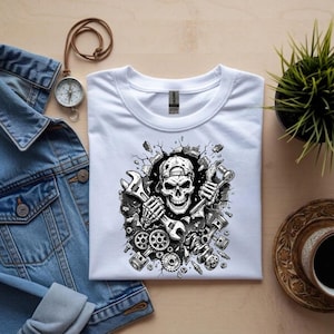 May include: White t-shirt featuring a black and white graphic of a skull wearing a cap and holding wrenches. The design includes gears, pistons, and a cracked wall effect, suggesting a mechanic theme.