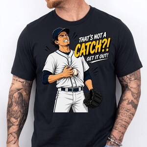 May include: Black t-shirt featuring a baseball player with a pained expression, holding a baseball. The text "THAT'S NOT A CATCH?! GET IT OUT!" is printed in yellow and white. The player is wearing a baseball uniform and a glove.