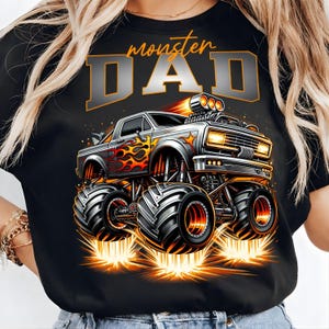 Pode incluir: T-shirt preto com um design de monster truck, chamas e o texto "monster DAD" em laranja e prata. O caminh&atilde;o tem pneus grandes e emite fa&iacute;scas. Um design divertido para um pai.