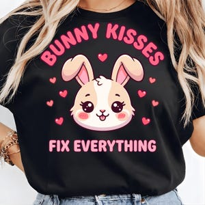 Könnte beinhalten: Schwarzes T-Shirt mit einem Cartoon-Hasen-Design. Der Text "BUNNY KISSES" wölbt sich über dem Hasen, darunter steht "FIX EVERYTHING". Der Hase ist hellbraun und weiß mit rosa Details, umgeben von rosa Herzen.