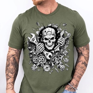 May include: Olive green t-shirt featuring a detailed graphic of a skull wearing a backwards baseball cap, holding wrenches. The skull is breaking through a wall, surrounded by engine parts. A unique design for mechanics and automotive enthusiasts.