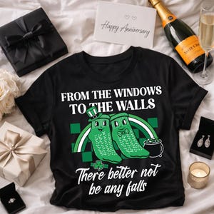 Op de afbeelding: Zwart t-shirt met de tekst "FROM THE WINDOWS TO THE WALLS" en "There better not be any falls". Het shirt heeft een afbeelding van twee groene sokken met een regenboog en een pot goud.