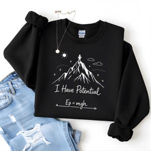 Puede incluir: Sudadera negra con un gráfico blanco de una montaña y una persona en la cima. El texto "I Have Potential" y una ecuación física están debajo. Un collar plateado y unos vaqueros azules también están en la imagen.