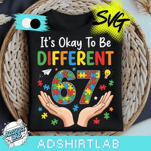 May include: Black t-shirt with the slogan "It's Okay To Be DIFFERENT" in various colours. The number "67" is made of puzzle pieces, with hands, a paper aeroplane, and a lightbulb. The shirt is displayed in a woven basket.