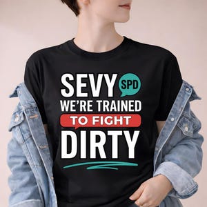 以下が含まれることがあります： 「SEVY WE'RE TRAINED TO FIGHT DIRTY」の文字が白、赤、ティールでプリントされた黒いTシャツ。クルーネックで、デニムジャケットと合わせて着用されています。