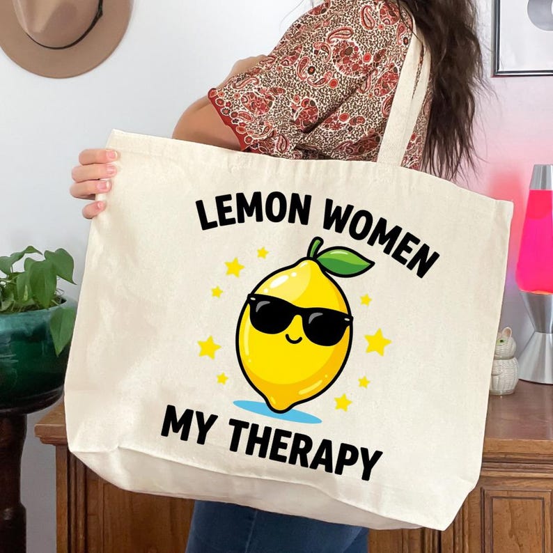 May include: Cream-colored tote bag with the text "Lemon Women My Therapy" and a cartoon lemon wearing sunglasses. The lemon is yellow with a green leaf and is surrounded by yellow stars. The bag has two cream-colored straps.
