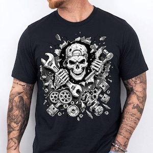 May include: Black t-shirt featuring a detailed graphic of a skull wearing a cap and holding wrenches. The skull is breaking through a wall, surrounded by engine parts like gears, pistons, and a crankshaft. The design is in shades of gray and white.