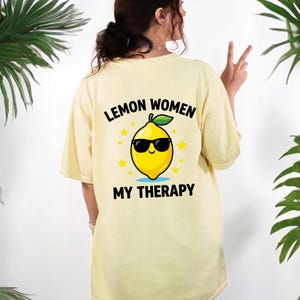 May include: A light yellow t-shirt with the text "LEMON WOMEN MY THERAPY" and a cartoon lemon wearing sunglasses. The lemon is yellow with a green leaf and is surrounded by yellow stars.