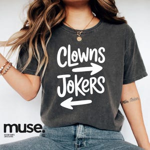 Op de afbeelding: Donkergrijs t-shirt met de witte tekst "Clowns Jokers" en pijlen. Dit casual t-shirt is geschikt voor dagelijks gebruik. Het merk "muse" staat onderaan het shirt.