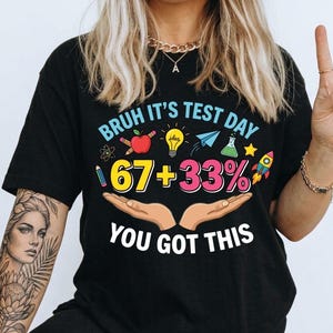 Może przedstawiać: Czarny t-shirt z napisem "Bruh It's Test Day" i równaniem "67 + 33%" w kolorowym wzorze. Koszulka zawiera ilustracje jabłka, żarówki, papierowego samolotu i rakiety. Na koszulce wydrukowano również słowa "You Got This".