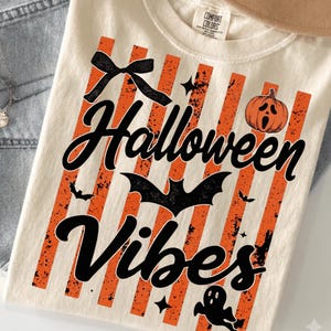 May include: A cream-colored t-shirt featuring a graphic print with vertical orange distressed stripes. The design includes the black text "Halloween VIBES" in a script font, a black bow, a black bat, a small ghost, and an orange screaming pumpkin.