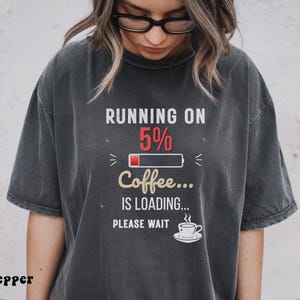 Puede incluir: Camiseta gris oscuro con el texto "RUNNING ON 5% Coffee... IS LOADING... PLEASE WAIT." El gráfico incluye un icono de batería y una taza de café. La camiseta es una prenda informal.