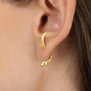Könnte beinhalten: Ein Paar goldfarbene Schlangenohrringe. Die Ohrringe sind so gestaltet, dass sie sich um das Ohr legen, wobei sich der Schlangenkopf unten und der Körper nach oben krümmt. Die Ohrringe haben eine strukturierte Oberfläche.