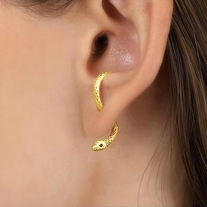 Puede incluir: Un par de pendientes dorados con forma de serpiente. Los pendientes se llevan en la oreja, con la cabeza de la serpiente en la parte inferior y el cuerpo curvándose hacia arriba. Los pendientes tienen un patrón texturizado y un pequeño acento rojo en la cabeza de la serpiente.