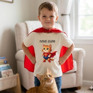Op de afbeelding: Een kind draagt een crèmekleurig t-shirt met de tekst "FUTURE LEGEND" en een superheldenkat. Het kind draagt ook een rode cape. Een oranje tabby kat staat op de voorgrond.