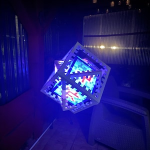 Op de afbeelding: Een geometrische, kubusvormige lamp met een pixelachtig ontwerp, verlicht met blauwe, rode en witte lichten. De lamp hangt in een donkere buitenomgeving, met een terrasstoel zichtbaar op de achtergrond.