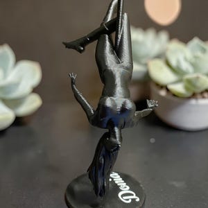 May include: A black 3D-printed figurine of a person pole dancing, upside down. The figure is on a black circular base with the name "Denia" in white script. The background includes a blurred succulent plant and a soft, round light.