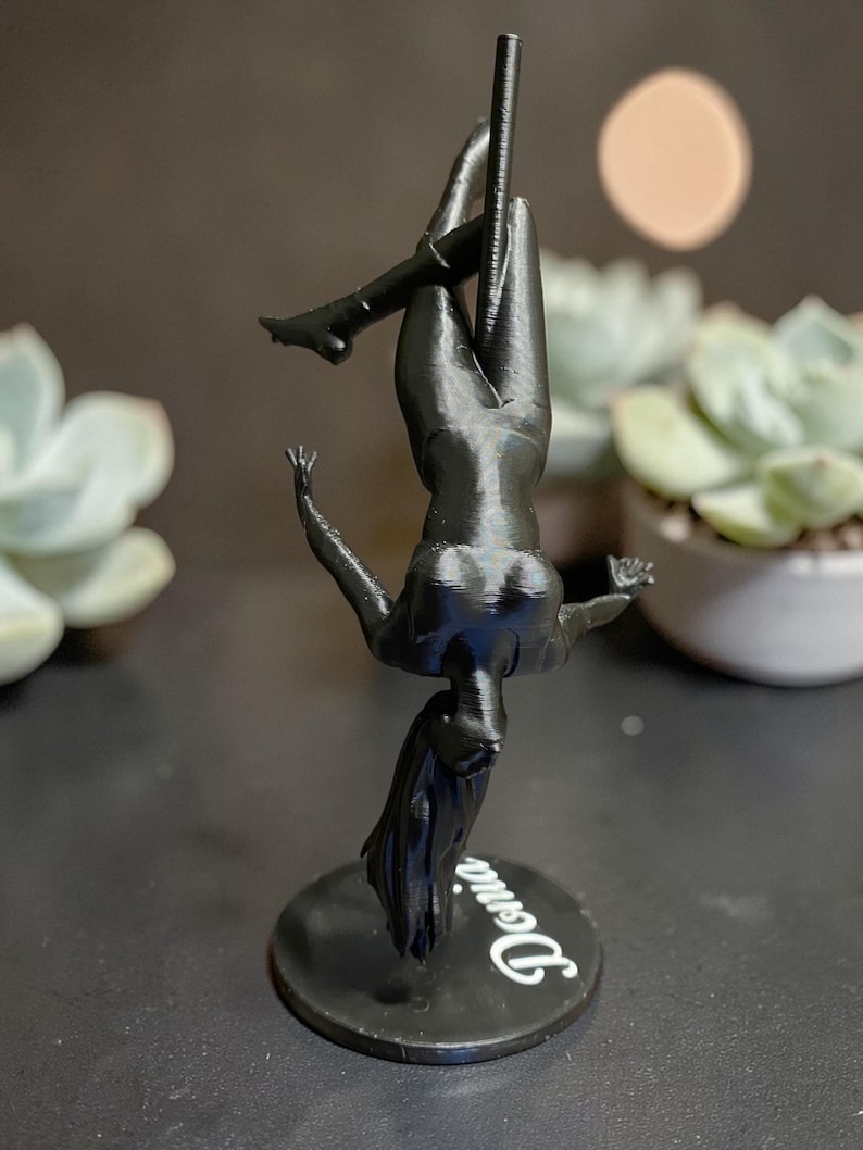 May include: A black 3D-printed figurine of a person pole dancing, upside down. The figure is on a black circular base with the name "Denia" in white script. The background includes a blurred succulent plant and a soft, round light.