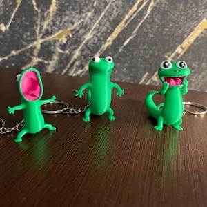Puede incluir: Tres llaveros de lagarto verde con expresiones divertidas. Uno tiene la boca abierta con el interior rosa, otro está de pie con los brazos extendidos y el tercero tiene una expresión de sorpresa. Cada llavero tiene un anillo plateado.
