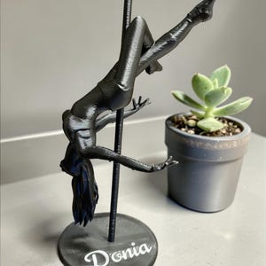 Könnte beinhalten: Eine schwarze 3D-gedruckte Skulptur einer Person, die Pole-Dance macht, mit dem Namen "Donia" auf der Basis. Die Figur hängt an einer Stange, mit ausgestreckten Armen und Beinen. Eine kleine Topf-Sukkulente befindet sich im Hintergrund.