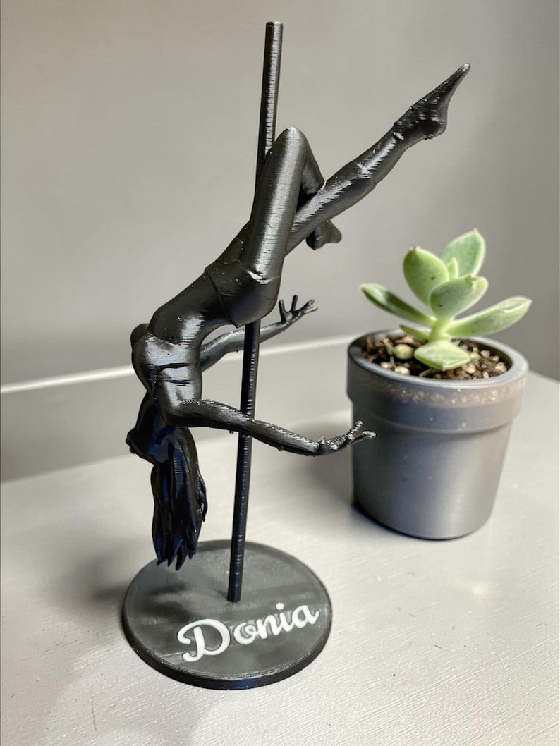 May include: A black 3D-printed sculpture of a person pole dancing, with the name "Donia" on the base. The figure is suspended from a pole, with arms and legs extended. A small potted succulent is in the background.