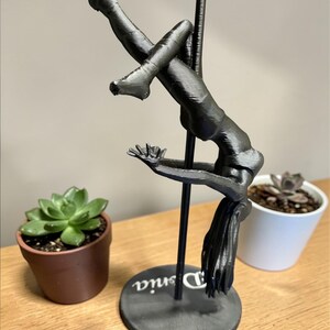 May include: A black sculpture of a person pole dancing, with the name "Denia" on the base. The figure is suspended from a black pole. Two potted succulents are on either side of the sculpture.