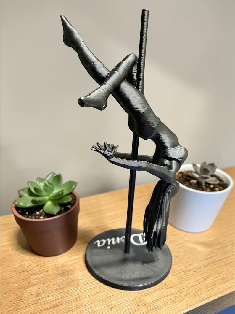 May include: A black sculpture of a person pole dancing, with the name "Denia" on the base. The figure is suspended from a black pole. Two potted succulents are on either side of the sculpture.