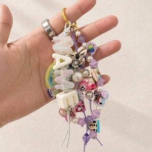 May include: A decorative keychain featuring a gold-tone ring, white block letters spelling NAME, and multiple strands of colorful beads. The beads include various shapes, sizes, and cartoon character motifs in shades of purple, pink, and white.