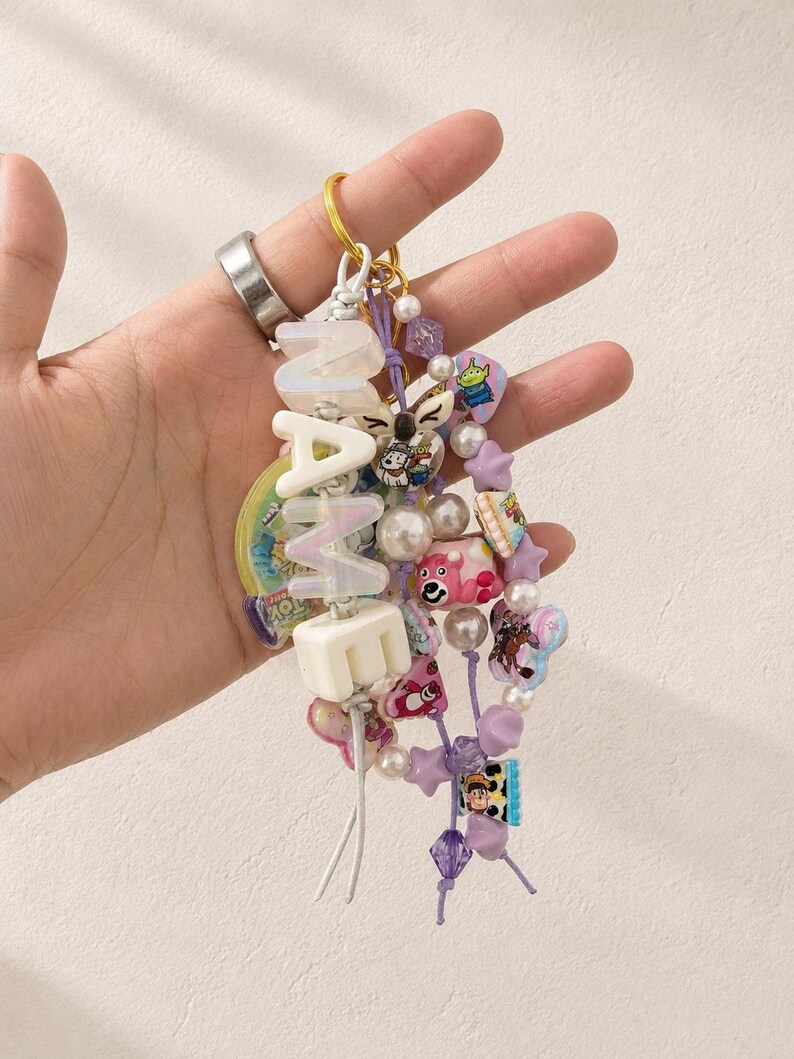 May include: A decorative keychain featuring a gold-tone ring, white block letters spelling NAME, and multiple strands of colorful beads. The beads include various shapes, sizes, and cartoon character motifs in shades of purple, pink, and white.