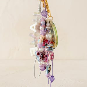 May include: A decorative keychain featuring a gold-toned ring and clasp, adorned with a variety of colorful beads including white, purple, and pink star-shaped, round, and bear-shaped charms, along with white letter beads spelling out MAMA.