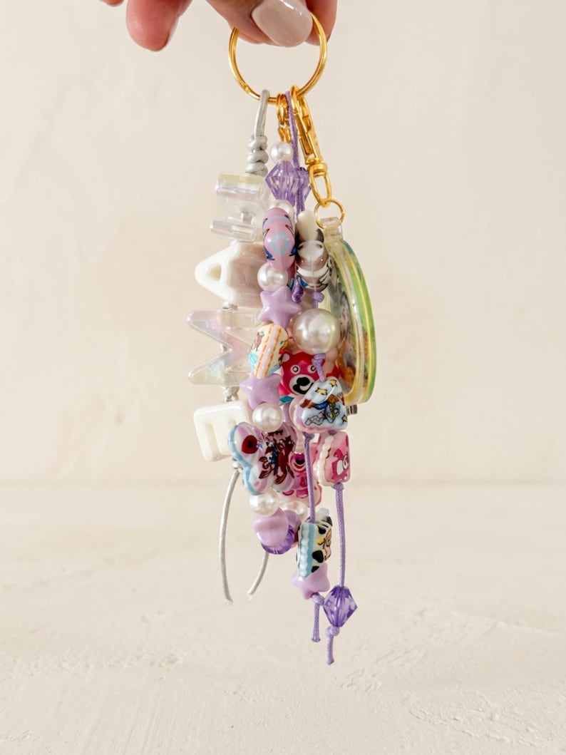 May include: A decorative keychain featuring a gold-toned ring and clasp, adorned with a variety of colorful beads including white, purple, and pink star-shaped, round, and bear-shaped charms, along with white letter beads spelling out MAMA.