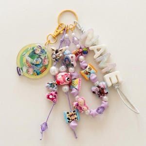 May include: A keychain featuring a gold-tone ring and clasp, a circular Toy Story character charm, and multiple strands of purple cord adorned with various colorful character beads, white pearls, star-shaped beads, and white letter beads spelling NAME.