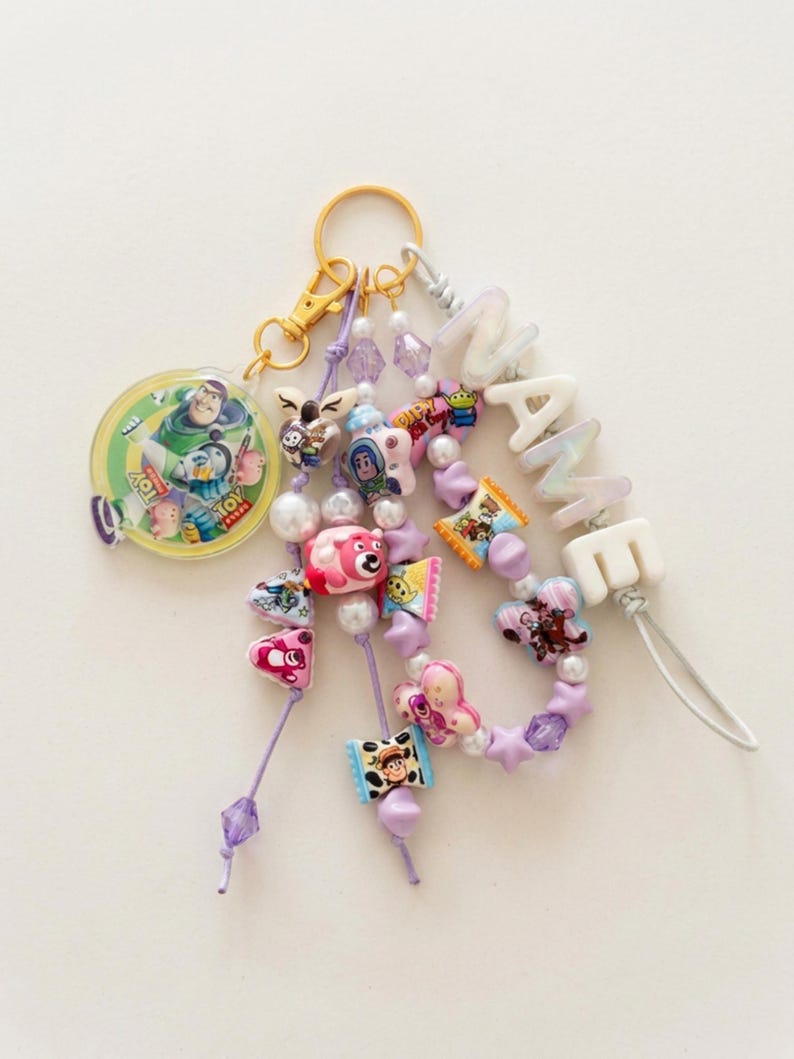 May include: A keychain featuring a gold-tone ring and clasp, a circular Toy Story character charm, and multiple strands of purple cord adorned with various colorful character beads, white pearls, star-shaped beads, and white letter beads spelling NAME.