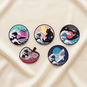 Puede incluir: Conjunto de cinco pines redondos de esmalte con diseños de inspiración japonesa. Cada pin muestra una imagen diferente: una ola con una montaña, un gato, un sol rojo, un cuenco de ramen y un tiburón. Los pines tienen bordes negros y colores vibrantes.