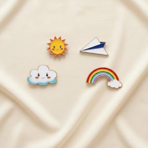 May include: Four enamel pins featuring cheerful designs. The pins include a smiling sun, a paper airplane, a cloud with a smiling face, and a rainbow with a white cloud. The pins are set against a cream-colored background.