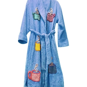 May include: A light blue terry cloth robe with a matching belt. The robe is decorated with embroidered coffee cup designs in various colors, including green, red, yellow, and blue. The robe has long sleeves and a shawl collar.