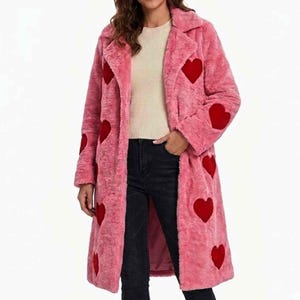 May include: A long, pink faux fur coat featuring a notched lapel collar and a pattern of dark red heart-shaped patches distributed across the front and sleeves. The coat is worn over a beige sweater and dark denim jeans, reaching below the hips.