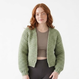 May include: A light green, open-front cardigan with a ribbed knit design. The cropped sweater has long sleeves and is worn over a brown top and dark gray leggings. The background is plain white.