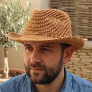 Könnte beinhalten: Ein hellbrauner Häkelhut mit Krempe im Fedora-Stil. Der Hut besteht aus einem strukturierten, gewebten Material und hat eine leicht gebogene Krempe. Der Hut hat eine neutrale Farbe und ist für verschiedene Anlässe geeignet.