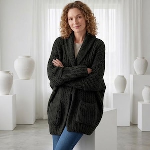 May include: A dark green, chunky knit cardigan with a shawl collar and two large front pockets. The cardigan is worn open, revealing a light-colored top underneath. The model is wearing blue jeans. The background features white vases and pedestals.