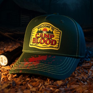 Peut inclure: Casquette de baseball vert foncé avec le texte "WELCOME TO CAMP BLOOD" en jaune et rouge. La casquette présente une éclaboussure de sang sur le bord. Une lampe de poche est en arrière-plan.
