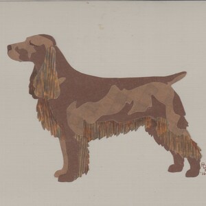 May include: A brown and tan paper cutout of a dog, possibly a Springer Spaniel, with a fringed mane. The dog is standing with its head turned to the left. The artist's signature and the year '16' are visible in the bottom right corner.