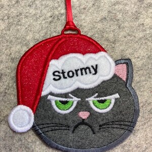 May include: A gray felt Christmas ornament shaped like a cat wearing a red Santa hat with white trim. The cat has green eyes, a pink nose, and a black mustache. The hat has the name "Stormy" embroidered on it.