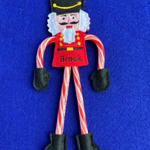 May include: A handmade Christmas ornament in the shape of a nutcracker soldier. The ornament features a red uniform with black boots and gloves, a black hat, and a white beard. The name "Brock" is printed on the uniform. The arms and legs are made of red and white striped candy canes.