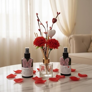 Puede incluir: Una exhibición temática de San Valentín con dos botellas de spray negras con etiquetas de mariposas y pétalos de rosa rojos. Un difusor de vidrio transparente con flores rojas y adornos de corazón está en el centro. Las botellas dicen "Valentine's Day" y "You & Me."