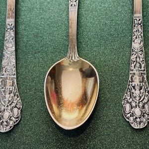 Könnte beinhalten: Drei verzierte Silberlöffel mit detaillierten floralen und architektonischen Designs. Die Löffel haben eine polierte goldene Schale und aufwendige Muster auf den Griffen. Sie sind auf einer dunkelgrünen Oberfläche angeordnet.