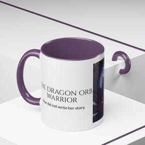 May include: A white ceramic mug with a purple interior and handle. The exterior features the text THE DRAGON ORB WARRIOR and Fear did not write her story. in black lettering, alongside a dark, detailed graphic illustration of a creature.