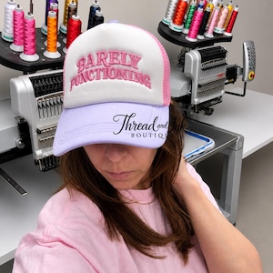 May include: A white and pink trucker hat with the embroidered text "Barely Functioning" in pink. The hat has a lavender brim and is part of a clothing and accessories line.