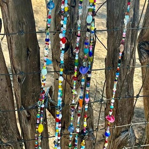 May include: Strands of colorful beads hang against a rustic wooden fence. The beads are a mix of shapes, sizes, and colors, including red, blue, yellow, and green. Some strands have small bells at the bottom.
