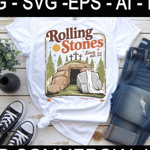 May include: A white t-shirt with the text "Rolling Stones Since ad 33" and a tomb illustration. The shirt is styled with blue jeans and black trainers. A small plant in a black vase and a pair of black trainers are also visible.